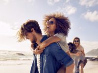 Group of friends walking along the beach, with men giving piggyback ride to girlfriends. Happy young friends enjoying a day at beach.